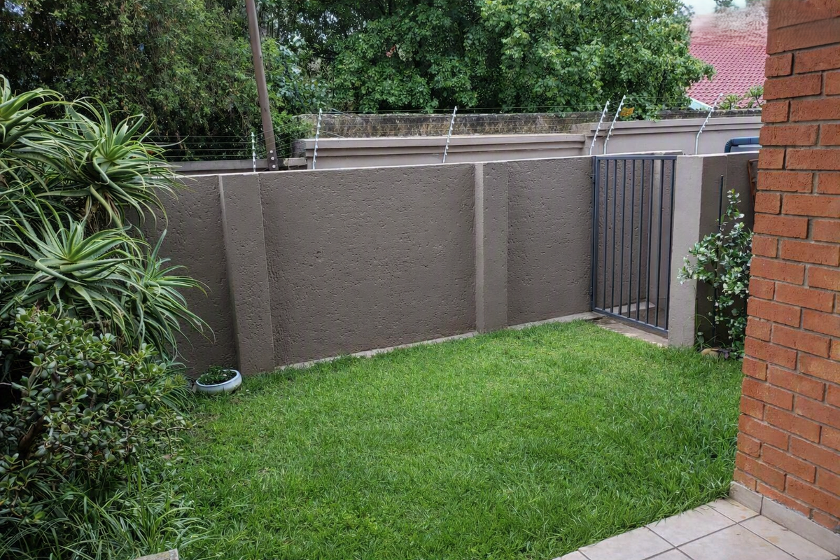 A private rear garden area featuring a well-maintained lawn, tiled patio, and secure boundary wall with electric fencing and a metal pedestrian gate.