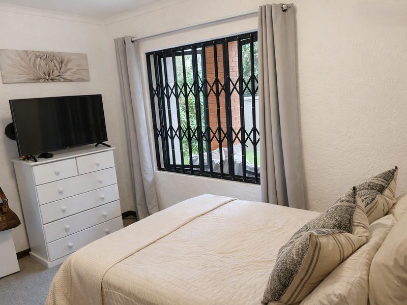A well-maintained bedroom featuring neutral walls, carpeted flooring, and a window fitted with a black security gate.