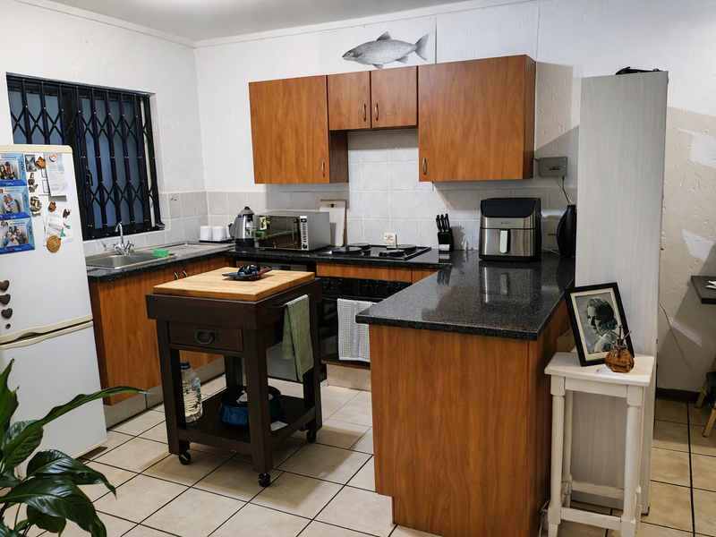 A functional kitchen featuring wood-look laminate cabinetry, dark granite countertops, ceramic tile backsplash, and tiled flooring with a window security gate.