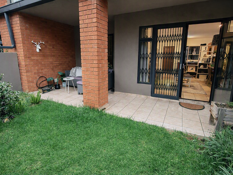A covered patio area with tiled flooring and a brick pillar, leading into the apartment through a security gate and sliding glass door.