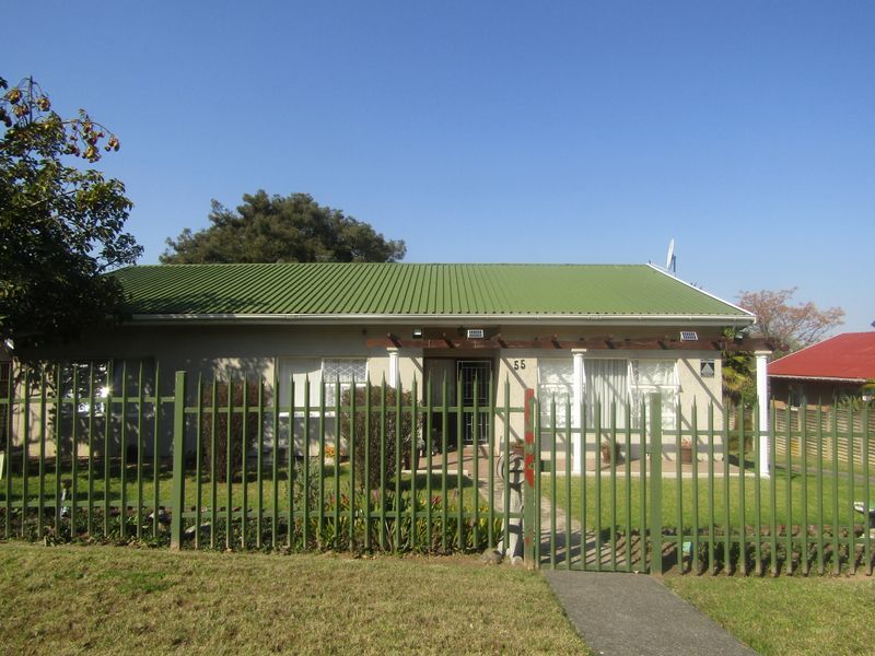 Front elevation of a single-story house featuring a green corrugated metal roof, timber pergola, and secure perimeter fencing with a gated entrance.