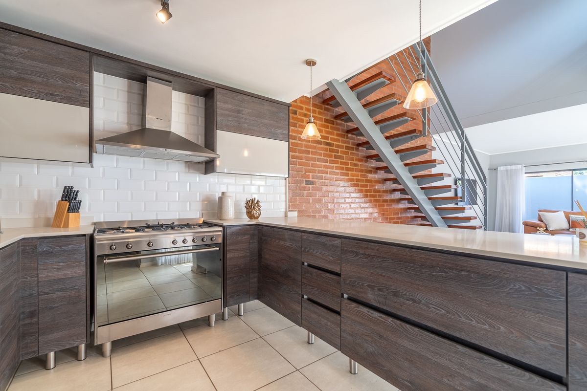 Modern kitchen featuring dark wood cabinetry, stone countertops, subway tile backsplash, and a stainless steel gas stove, adjacent to an exposed brick staircase.