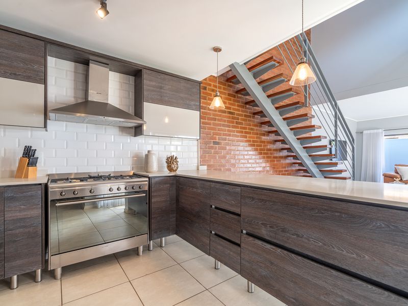 Modern kitchen featuring dark wood cabinetry, stone countertops, subway tile backsplash, and a stainless steel gas stove, adjacent to an exposed brick staircase.