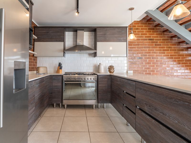 Modern kitchen featuring dark wood cabinetry, stone countertops, subway tile backsplash, and an exposed brick wall, adjacent to an open staircase.