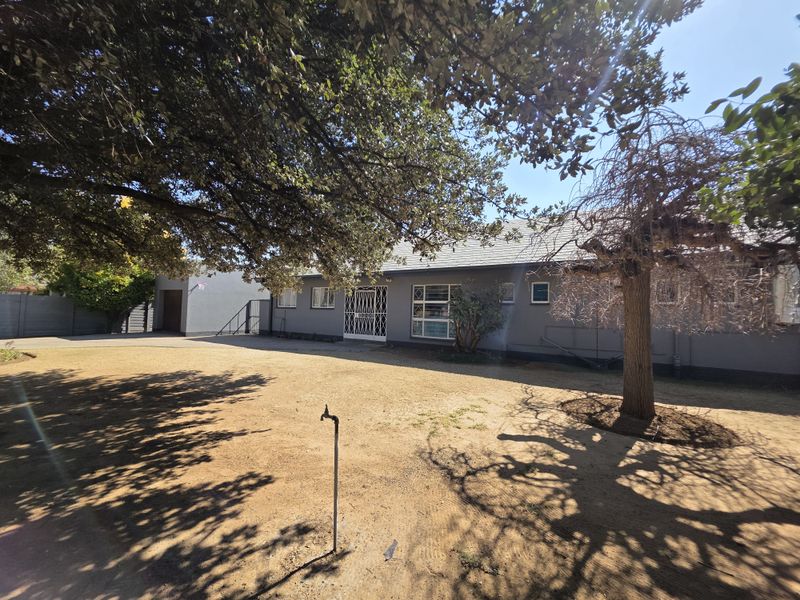 Front view of a single-story house featuring a tiled roof, security gate, and attached garage, set on a large plot with mature trees.