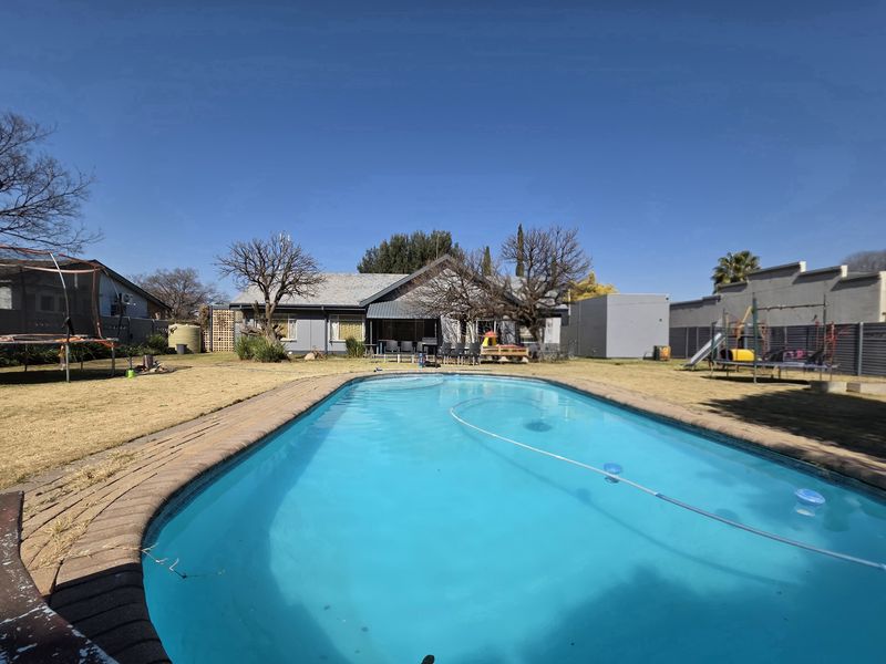 A spacious backyard featuring a swimming pool with brick coping, surrounded by a large lawn area and rear view of the house.