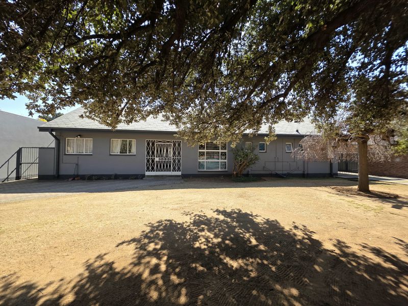 Front elevation of a single-story house featuring a paved driveway, security gates, and a large mature tree providing shade over the front yard.