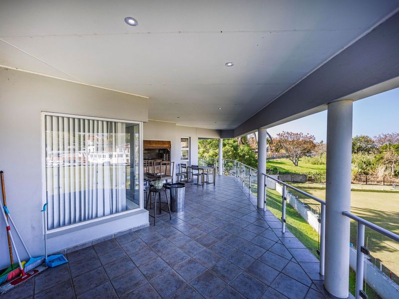 Spacious covered balcony featuring tiled flooring, a built-in braai, and a glass balustrade overlooking the surrounding landscape.