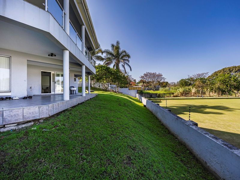 Rear view of the property featuring a covered tiled patio with glass balustrades, overlooking a landscaped garden and retaining wall with electric fencing.
