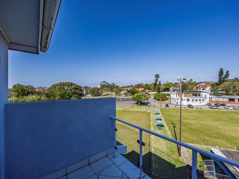A clean, elevated balcony featuring a modern glass balustrade and tiled flooring, offering an expansive view over the surrounding neighborhood and green space.