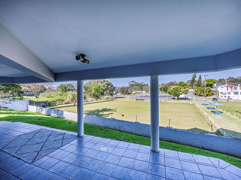 A spacious covered patio featuring tiled flooring and support columns, offering an open view overlooking a green field and surrounding neighborhood.