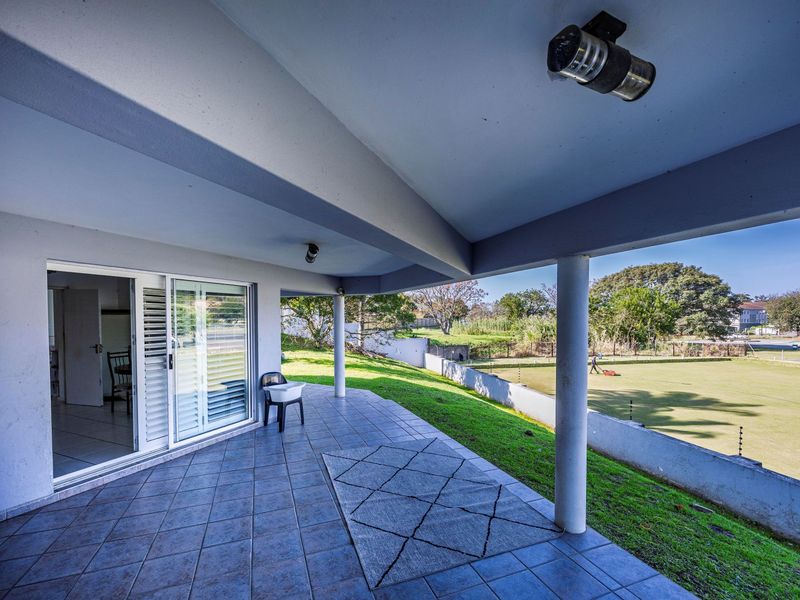 A spacious covered patio featuring tiled flooring, sliding glass doors with security shutters, and views overlooking a landscaped lawn and boundary wall.