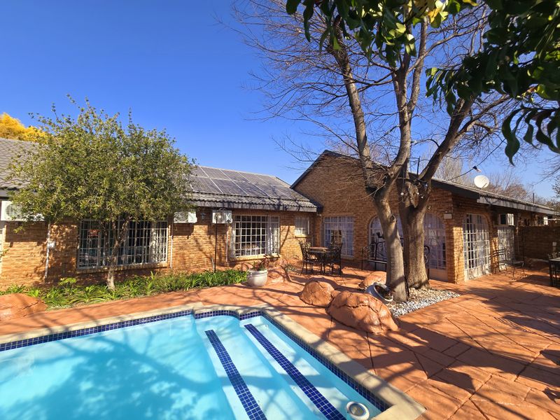 A well-maintained outdoor pool area featuring a paved patio, brick exterior walls, and roof-mounted solar panels.