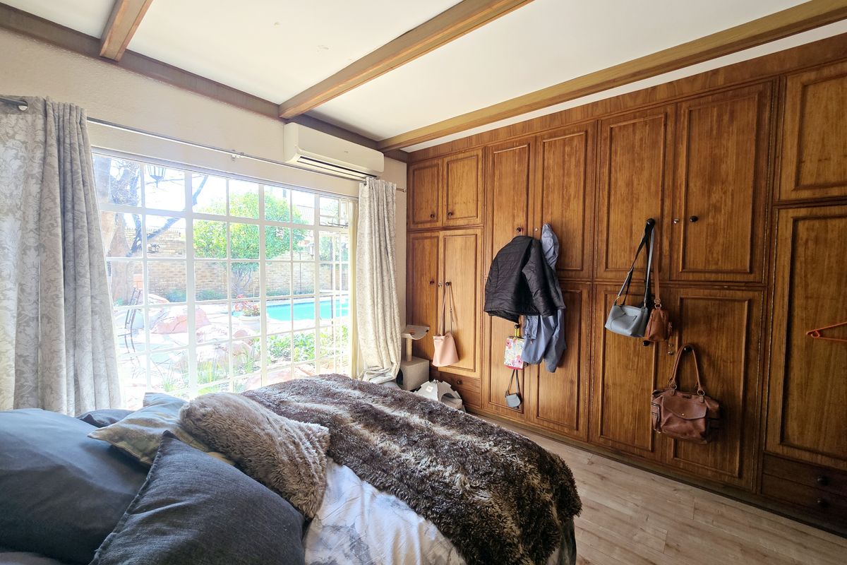 A bedroom featuring extensive built-in wooden cabinetry, exposed ceiling beams, laminate flooring, and a large window overlooking the garden and pool area.