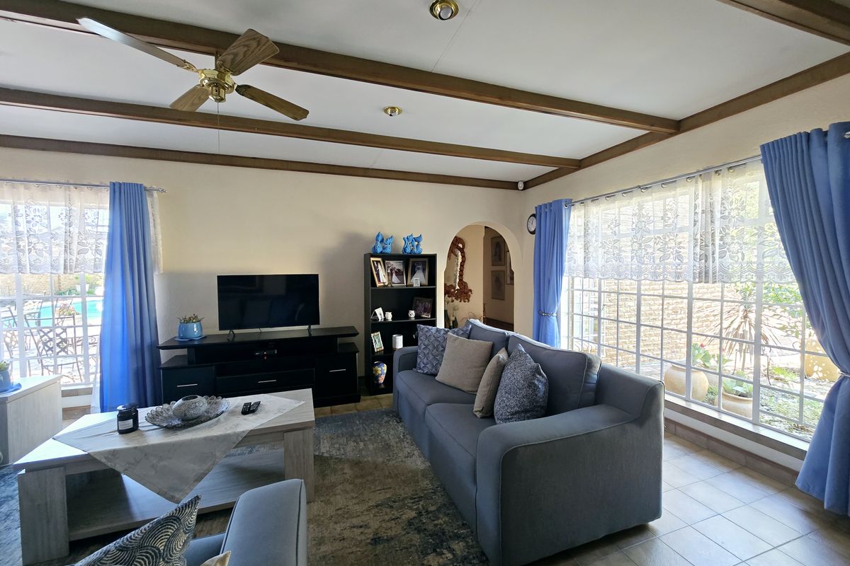 A comfortable living room featuring exposed ceiling beams, a ceiling fan, tiled flooring, and large windows that provide ample natural light.