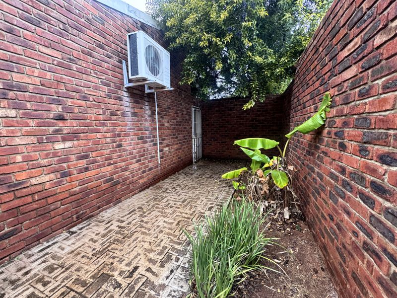 A narrow rear exterior passage featuring exposed brick walls, paved flooring, and a wall-mounted air conditioning unit.