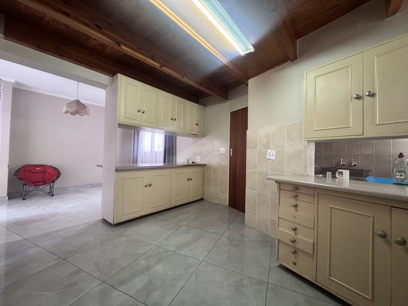 A functional kitchen featuring dated cabinetry, tiled splashbacks, and exposed timber ceiling beams, with an open-plan layout connecting to the adjacent living area.