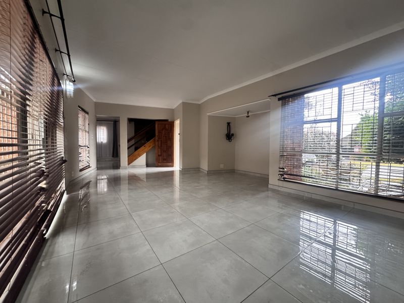 Spacious, light-filled living area featuring large porcelain floor tiles, a wooden staircase, and large windows with security bars and timber blinds.