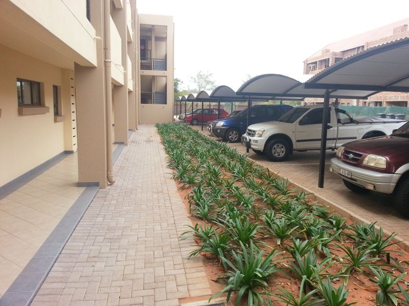 Exterior view of an apartment complex featuring a paved walkway, landscaped garden beds, and covered parking bays for residents.