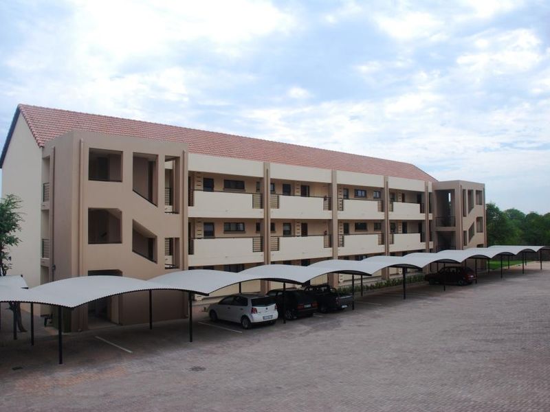 A multi-story apartment complex featuring a tiled roof, external stairwells, private balconies, and a large paved parking area with covered carports.