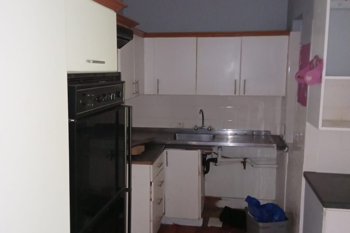 A functional kitchen featuring built-in cabinetry, a wall-mounted oven, and a stainless steel sink, currently requiring maintenance and cosmetic updates to the cabinetry and flooring.