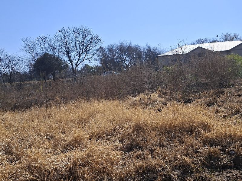 A view of a vacant residential plot featuring dry, overgrown grass and a boundary wall, with neighboring structures visible in the background.