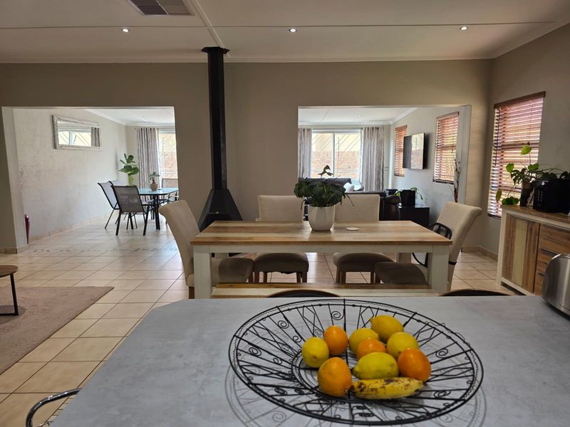 Spacious open-plan living and dining area featuring tiled floors, a central freestanding fireplace, and abundant natural light through large windows with venetian blinds.