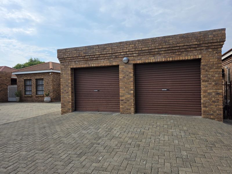 A double garage with brown roller doors and face brick facade, situated on a paved driveway within a townhouse complex.