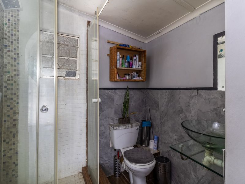 A compact bathroom featuring a glass shower enclosure with mosaic tiling, a glass vessel sink, and a toilet, requiring some cosmetic updates.