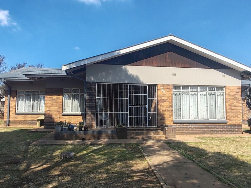 Front view of a single-story brick house featuring a metal roof, security gate entrance, and a spacious front garden with a paved walkway.
