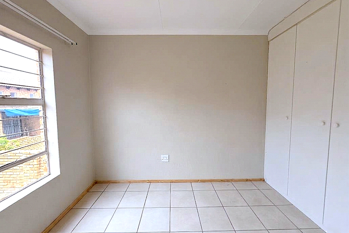 A bright, empty bedroom featuring neutral tiled flooring, built-in cupboards, and a large window providing natural light.