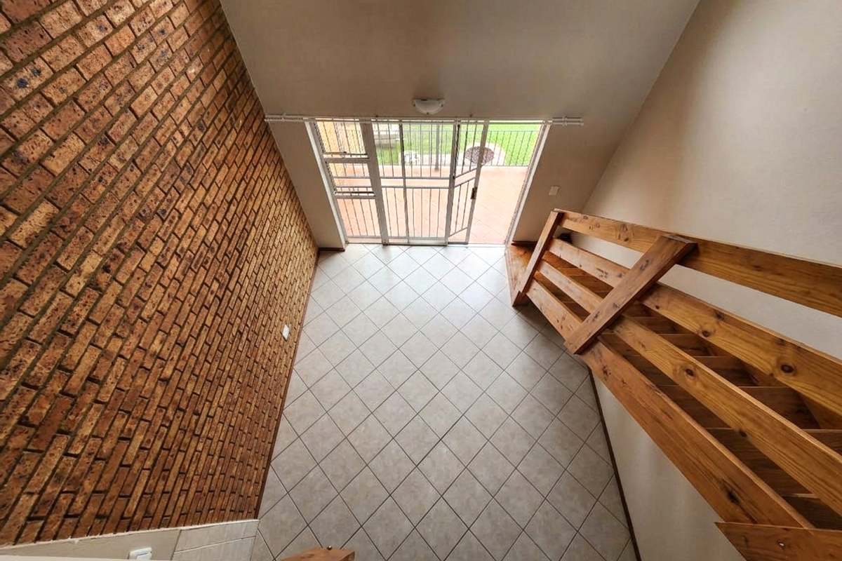 An empty living area featuring an exposed brick wall, tiled flooring, and a wooden staircase leading to an upper level with access to the patio.