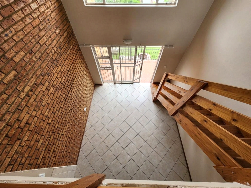 An empty living area featuring an exposed brick wall, tiled flooring, and a wooden staircase leading to an upper level with access to the patio.