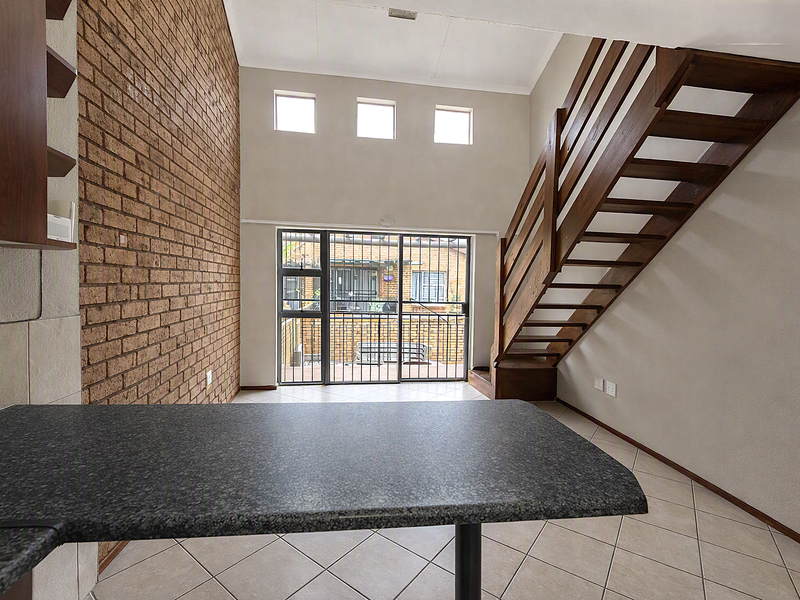 Open plan living area featuring a double-volume ceiling, exposed brick feature wall, wooden staircase, and direct access to the rear patio through sliding doors.
