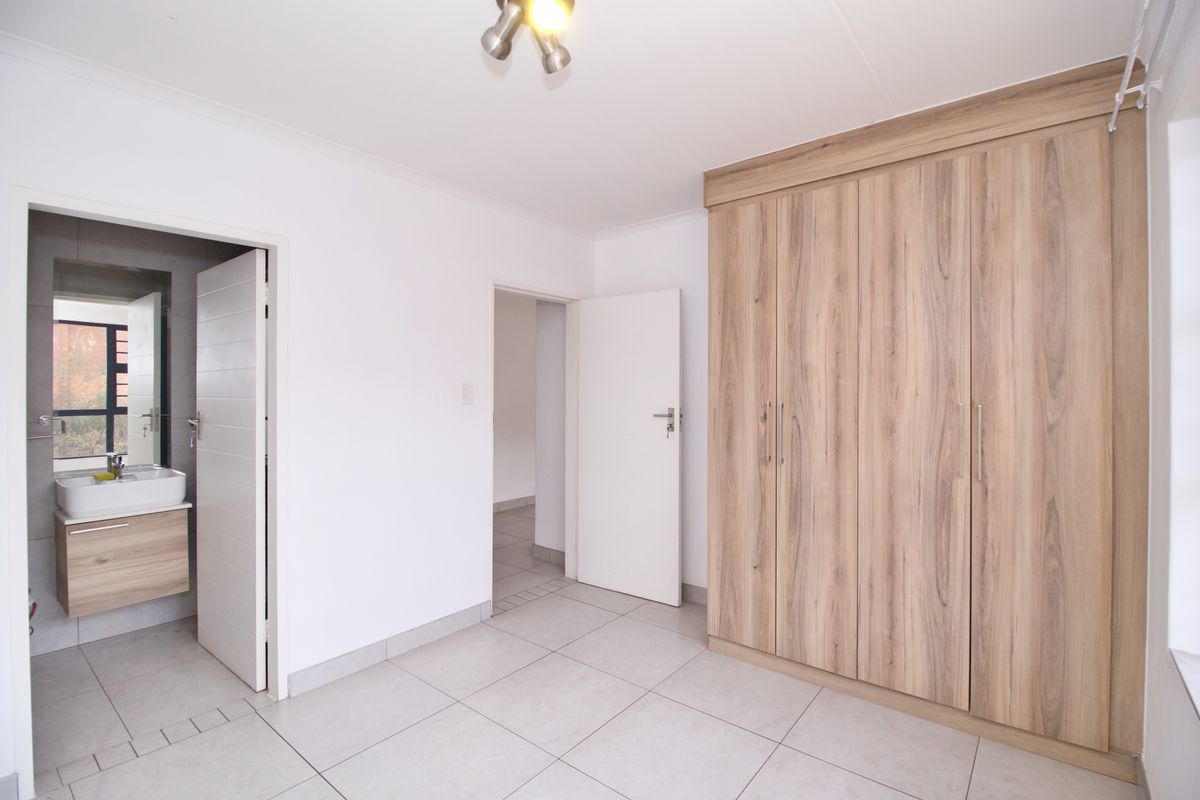 A bright, empty bedroom featuring modern wood-finish built-in cupboards, neutral tiled flooring, and direct access to an en-suite bathroom.