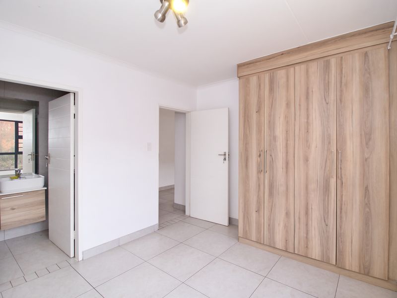 A bright, empty bedroom featuring modern wood-finish built-in cupboards, neutral tiled flooring, and direct access to an en-suite bathroom.