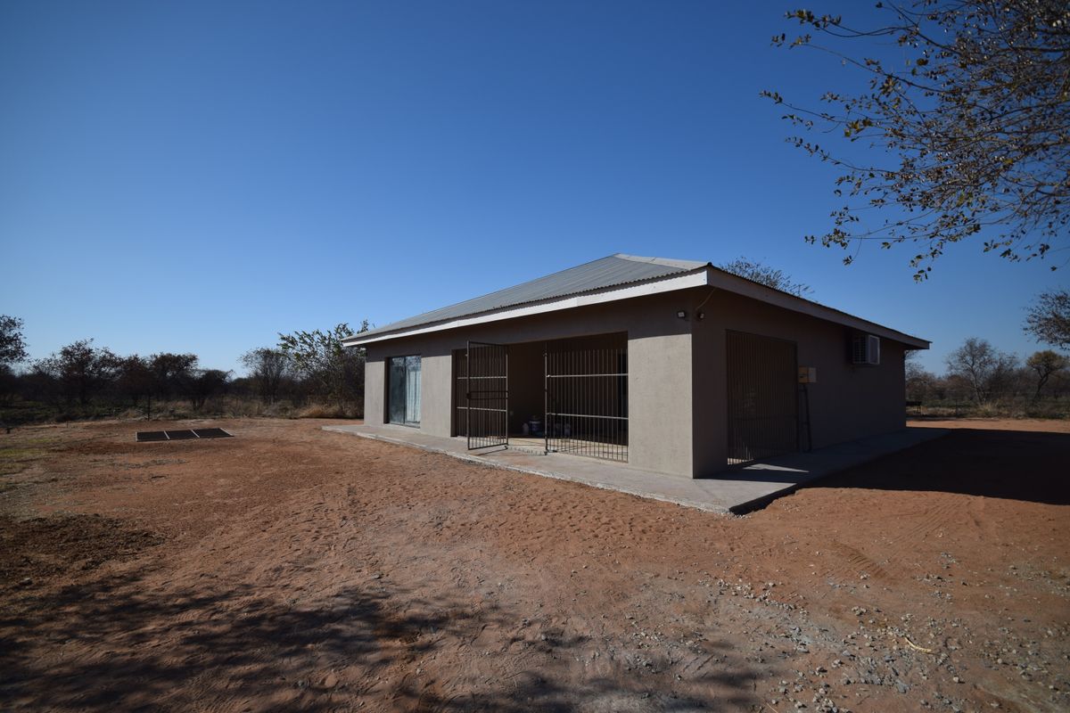 Rear view of a single-story house featuring a corrugated metal roof, security gates, and a concrete apron set in a rural landscape.