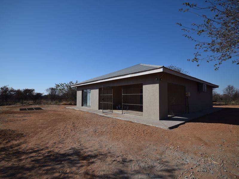 Rear view of a single-story house featuring a corrugated metal roof, security gates, and a concrete apron set in a rural landscape.
