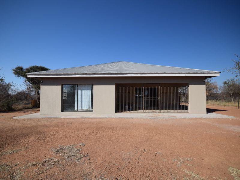 A single-story house featuring a corrugated metal roof, plastered exterior walls, a concrete patio, and secure metal gate access.