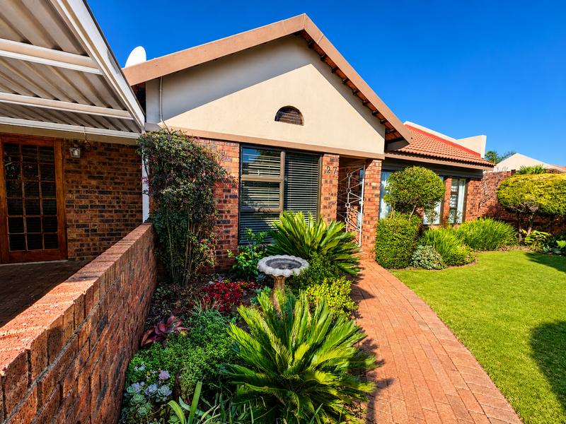 A well-maintained townhouse front elevation featuring a brick facade, paved walkway, covered patio area, and established garden landscaping.