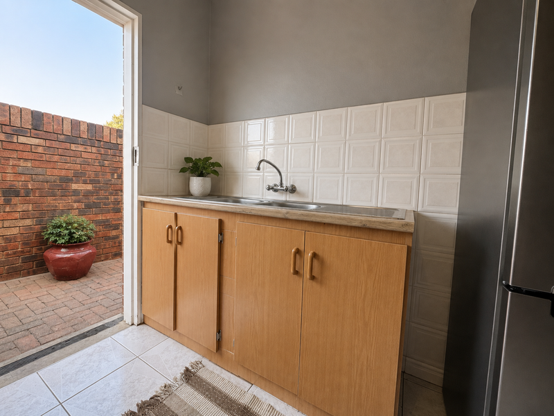 A functional scullery area featuring laminate cabinetry, a stainless steel sink, and a tiled backsplash, with direct access to an outdoor paved patio.