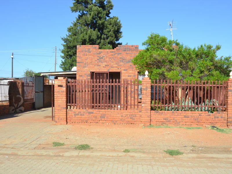Front view of a face brick house featuring a secure metal gate, perimeter brick wall with metal fencing, and a paved driveway.