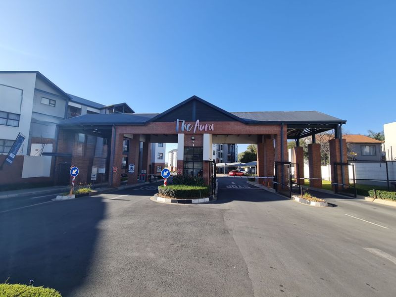 The main entrance to The Aura apartment complex, featuring a brick security gatehouse, boom gates, and paved driveway access.