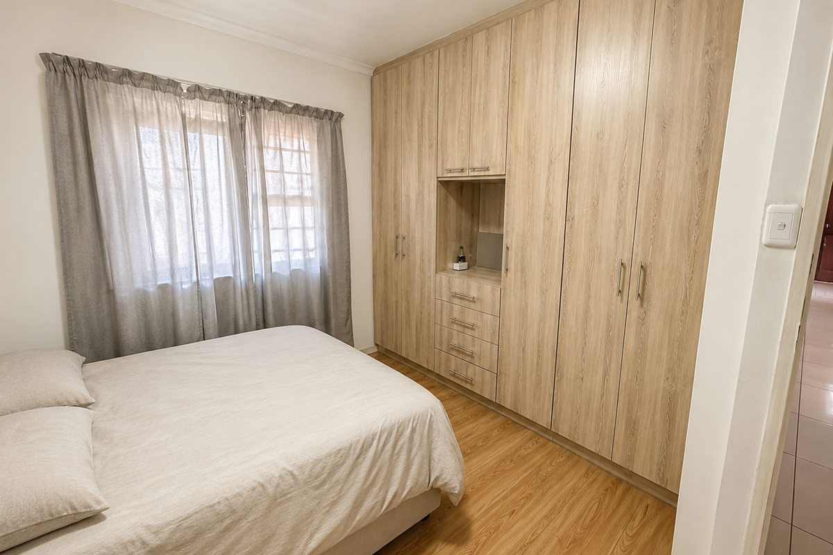 A well-maintained bedroom featuring light wood-grain built-in cupboards, laminate flooring, and a window with neutral curtains.