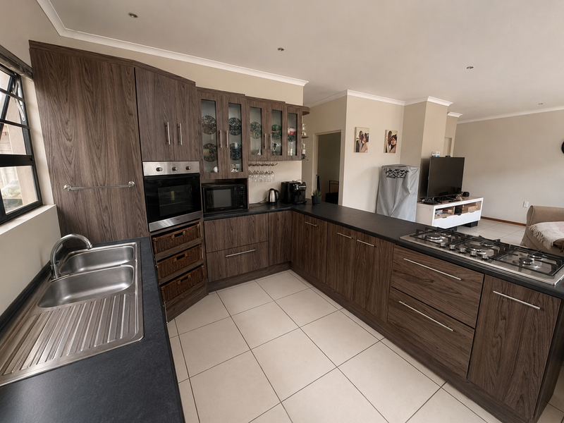 Modern open-plan kitchen featuring dark wood-grain cabinetry, stone-look countertops, a gas hob, built-in oven, and tiled flooring, seamlessly flowing into the living area.