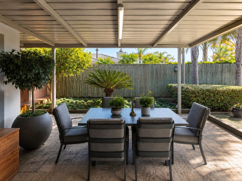 A well-maintained covered patio featuring paved flooring and a metal roof structure, overlooking a landscaped garden with bamboo fencing.