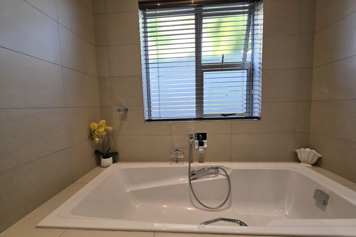 A modern bathroom featuring a built-in bathtub with chrome fixtures, neutral wall tiling, and a window with venetian blinds.