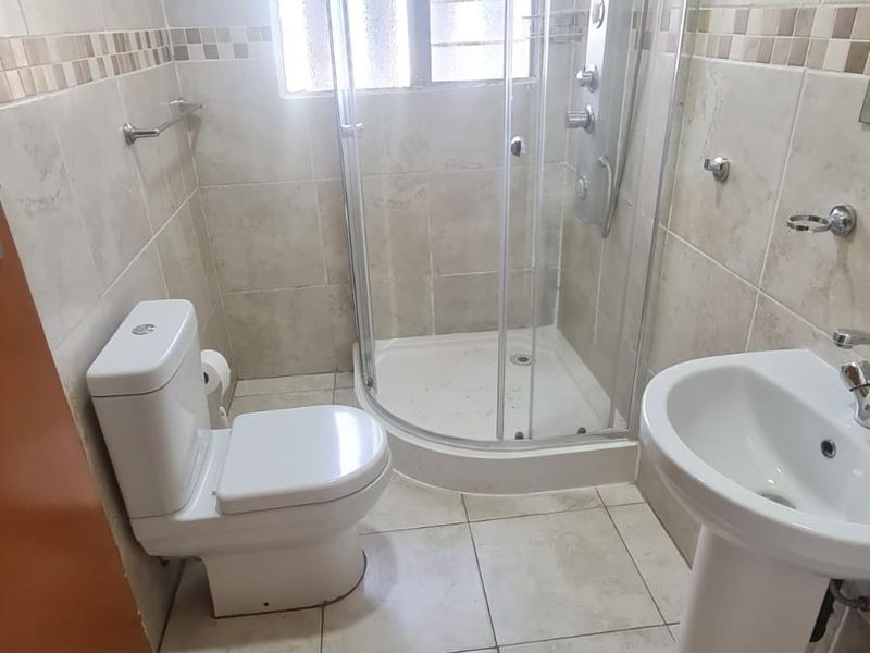 A functional bathroom featuring a corner shower enclosure, pedestal basin, and toilet with neutral wall and floor tiling.