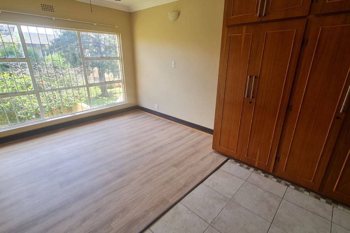 A bright, empty bedroom featuring built-in wooden cupboards, a mix of laminate and tiled flooring, and a large window with security bars.