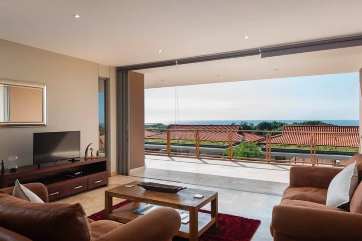 Spacious living room featuring large sliding glass doors that open onto a balcony with scenic coastal views and tiled flooring throughout.